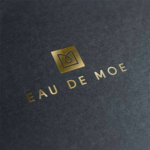 Eau De Moe text and emblem logo in shiny gold on a dark grey background